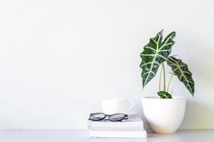 bril en witte koffiekop op het boek en in de buurt van alocasia sanderiana stier of alocasia plant op tafel en witte muur achtergrond foto