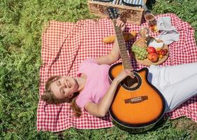 bovenaanzicht van jonge vrouw in witte broek buiten met picknick, eten en gitaar spelen foto