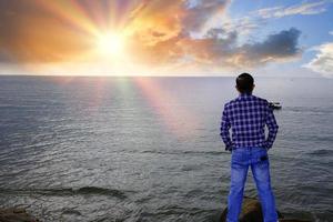 portret knappe man staande op een steen aan de kant van de zee en ver over de zee kijkend met een zonsopgang ochtend avontuur reis concept. foto