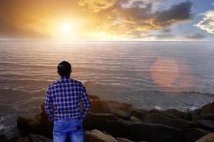 portret knappe man staande op een steen aan de kant van de zee en ver over de zee kijkend met een zonsopgang ochtend avontuur reis concept. foto