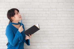 mooie Aziatische vrouw die lacht staande denken en schrijven notebook op betonnen cement witte achtergrond thuis, meisje huiswerk op boek, onderwijs en lifestyle concept. foto