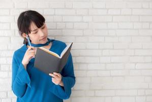 mooie Aziatische vrouw die lacht staande denken en schrijven notebook op betonnen cement witte achtergrond thuis, meisje huiswerk op boek, onderwijs en lifestyle concept. foto