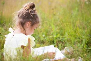 meisje in een gele jurk zit in het gras op een deken in een veld en leest een papieren boek. internationale kinderdag. zomertijd, jeugd, onderwijs en amusement, cottage core. kopieer ruimte foto
