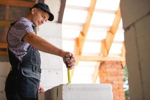 bouwvakker op bouwplaats meet de lengte van raamopening en bakstenen muur met meetlint. cottage zijn gemaakt van poreuze betonblokken, werkkleding - jumpsuit en baseballcap foto