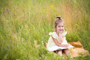 meisje in een gele jurk zit in het gras op een deken in een veld en leest een papieren boek. internationale kinderdag. zomertijd, jeugd, onderwijs en amusement, cottage core. kopieer ruimte foto