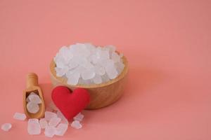 witte kandijsuiker, kristalsuiker in houten lepel en glazen pot met rood hart op roze achtergrond. geselecteerde focus. ruimte voor tekst. foto