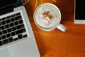 hete latte in beker op houten tafel uitzicht vanaf de top met laptop en smartphone rond en kopieer ruimte. foto