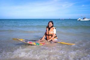 glimlach, gelukkige aziatische vrouw met levensreddend jasje zit op een klein paddleboard met roeispaan op het golvende zeestrand foto