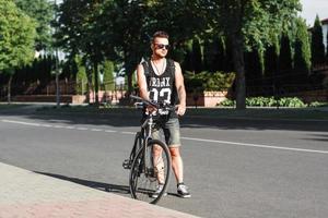 stijlvolle jongeman staande op de weg met de fiets op de achtergrond van groen. foto