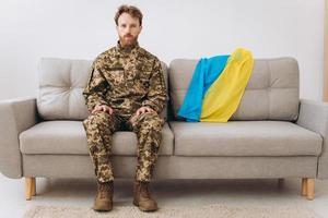 portret van een emotionele jonge Oekraïense patriot-soldaat in militair uniform zittend op kantoor op een bank met een gele en blauwe vlag. foto