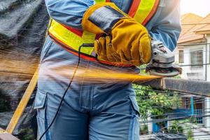 jonge man lasser in een werkende blauwe overall en werkhandschoenen maalt een metalen voorwerp met een haakse slijper in de garage, heldere vonken vliegen naar de zijkanten foto