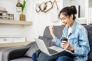 Aziatische tienervrouw die op een bank in het huis zit en koffie drinkt, geniet van het werk door de computerlaptop te gebruiken om geld te verdienen met online zaken, vanuit huis te werken foto