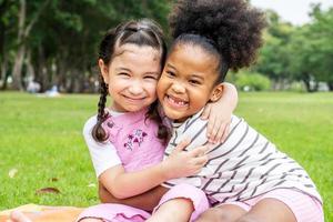 twee Afro-Amerikaanse vrolijke kleine meisjes zitten op de mat en knuffelen elkaar met liefde in de tuin foto