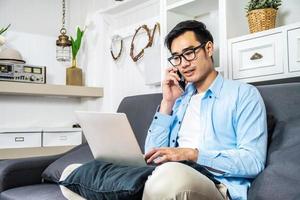 Aziatische zakenman zit op de bank in zijn eigen huis en belt op zijn mobiele telefoon om informatie te controleren die hij op zijn laptop heeft ontvangen met een ernstig bezorgde uitdrukking, werk vanuit huis foto