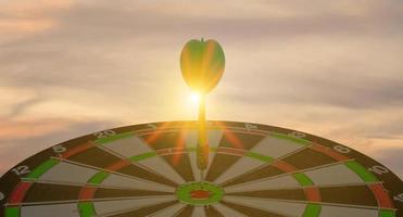 silhouet van groene dartpijl die het dartbord van het bullseye-doelcentrum op zonsondergangachtergrond raakt. bedrijfstargeting en focusconcept, strategie, prestatie en planningsconcept foto