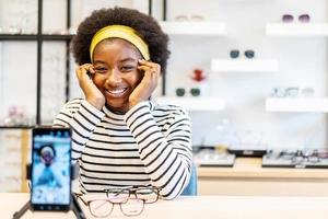 jonge vrouw Afro-Amerikaanse ondernemer is modevlogger die een bril op mobiele telefoon laat zien en glimlacht om het online te verkopen livestreaming in een brillenwinkel foto