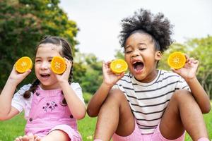 twee Afro-Amerikaanse kleine meisjes die op de mat zitten en sinaasappels eten in het herfstpark, spelen met sinaasappels foto