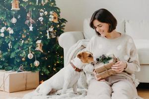 foto van blije brunette vrouw houdt kerstcadeau vast, poseert op de vloer met rashond jack russell terrier hond tegen gezellig interieur, versierde nieuwjaarsboom. feestelijke stemming, viering concept.