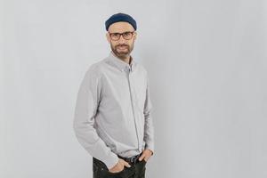 zijwaarts schot van knappe tevreden man draagt stijlvolle hoed, houdt handen in de zakken, staat tegen een witte achtergrond met kopieerruimte voor uw advertentie of promotie. hipster kerel modellen binnen foto