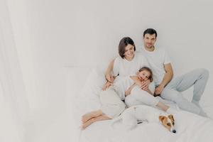 gelukkige familie pose op wit bed tijdens het weekend. vader, moeder, hun dochter en hond voelen zich op hun gemak in de slaapkamer, omhelzen en hebben plezier, ontspannen en vrolijk zijn. vriendelijke familie en rust foto