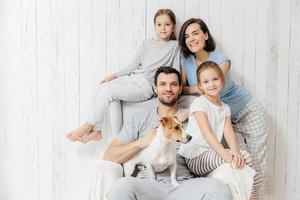 familie portret. gelukkige ouders met hun twee dochters en hond poseren samen tegen een witte achtergrond, brengen vrije tijd thuis door, in een goed humeur. moeder, vader en kleine zusjes poseren binnen foto