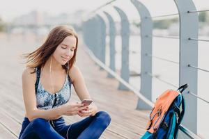 positief fitnessmeisje in top en legging, heeft een blije uitdrukking als een aangenaam sms-bericht van vriendje leest, heeft donker haar, rust buiten. mensen, sport, moderne technologieën en lifestyle foto