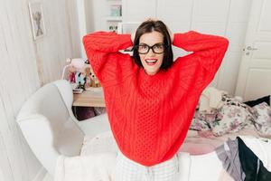 horizontaal portret van vrolijke brunette huisvrouw draagt rode warme trui en bril, poseert tegen het interieur in haar kamer, heeft een positieve glimlach. mooie vrouw met vrolijke uitdrukking binnen foto