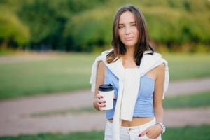 buiten schot van brunette vrouw gekleed in casual t-shirt, witte broek, houdt hand in zak, drinkt aromatische koffie tijdens ochtendwandeling, staat tegen groene natuur achtergrond. vrije tijd concept foto