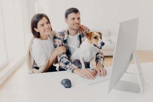 indoor shot van vrolijke man en vrouw kijken met vrolijke uitdrukkingen, lachen als kijken naar grappige film, rusten samen in vrije tijd, nieuwsgierige hond kijkt aandachtig naar monitor van computer, man toetsenborden foto