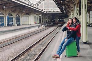 mensen, reizen, relatieconcept. gelukkige ongeschoren man en vrouw omhelzen elkaar op het treinstation, gaan op reis, hebben vakantie. knappe man spreekt op smartphone, ontvangt knuffel vriendin foto