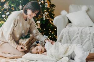 famaily, gelukkig wintervakantie concept. zorgzame moeder staat op knieën in de buurt van slapende dochter, bereidt cadeautjes voor op nieuwjaar, heeft een aangename glimlach, poseert in een gezellige woonkamer. kerstnacht en wonder foto