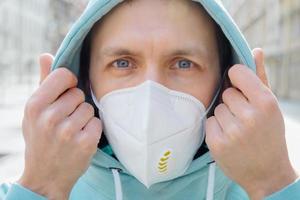 close-up shot van europese man beschermt gezicht met ademhalingsmasker, draagt hoodie, poseert buiten op straat, kijkt serieus naar camera, voorkomt coronavirus of covid-19, griep. straat tijdens quarantaine foto