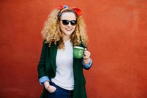 aantrekkelijke jonge Europese vrouw met hoofdband met een stijlvolle jas en tinten met een kopje cappuccino met een gelukkige uitdrukking terwijl ze tegen een oranje achtergrond staat. mensen, levensstijlconcept foto