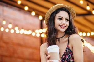 verlegen brunette vrouw met donkere ogen en rode lippen, gekleed in strohoed en jurk, met afhaalmaaltijden koffie of thee kijkend met een glimlach opzij, blij haar vriendje op te merken, rustend op het terras foto