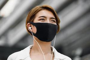 jonge aziatische vrouw die buiten gezichtsmasker draagt terwijl ze naar muziek luistert in de stad tijdens de covid-19 pandemie foto