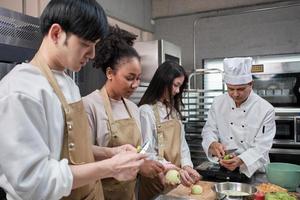 hobbykeukencursus, senior mannelijke chef-kok in kookuniform leert jonge kooklesstudenten appels te schillen en hakken, ingrediënten voor banketbakkerswaren, fruittaarten in de roestvrijstalen keuken van het restaurant. foto