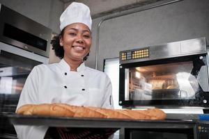 portret van een Afro-Amerikaanse vrouwelijke chef-kok in een wit kookuniform die naar de camera kijkt met een vrolijke glimlach en trots met een dienblad met stokbrood in de keuken, professionele banketbakkers, verse bakkerijbezetting. foto