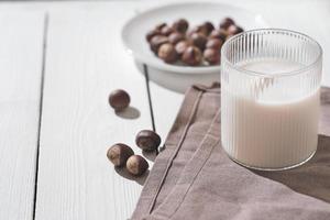 zuivelvervanging, gezonde plantaardige melk in een glas. hazelnootdrank op een witte houten tafel naast noten op een bord.. foto