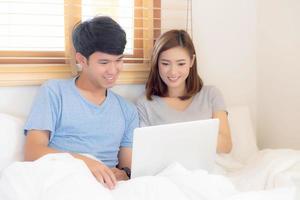 mooie jonge Aziatische paar glimlach surfen op internet samen kijken naar film online op bed met laptopcomputer om samen te ontspannen, Azië vrouw en man werken notebook in slaapkamer, lifestyle concept. foto