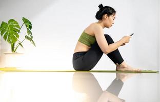 vrouwen die mobiel spelen en op de groene yogamat zitten terwijl ze uitrusten voor een doe-oefening in huis. foto