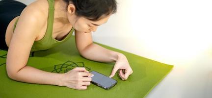 vrouwen die mobiel spelen en op de groene yogamat zitten terwijl ze uitrusten voor een doe-oefening in huis. foto