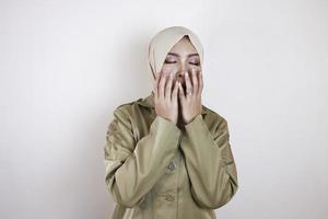 portret van slaperige aziatische moslimvrouw, gekleed in een bruin uniform en hijab, die haar mond bedekt met een hand terwijl ze geeuwt. foto