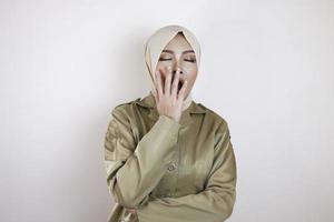 portret van slaperige aziatische moslimvrouw, gekleed in een bruin uniform en hijab, die haar mond bedekt met een hand terwijl ze geeuwt. foto