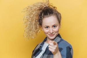 positieve zelfverzekerde mooie vrouw met krullende paardenstaart, geeft direct op camera aan, kiest jou, gekleed in casual kleding, geïsoleerd op gele achtergrond. shot van mooie vrouwengebaren binnen foto