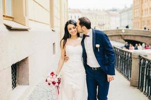 aanhankelijk knappe bruidegom met baard kust zijn toekomstige vrouw. mooie brunette bruid draagt mooie jurk en houdt bouquete vast, loopt met minnaar op brug. huwelijk en viering concept foto