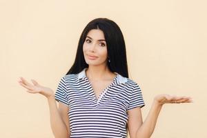 shot van aarzelende mooie vrouw haalt schouders op met verbazing, weet geen antwoord op moeilijke vraag, poseert tegen lichtroze achtergrond. onwetende brunette vrouw heeft twijfelachtige uitdrukking foto