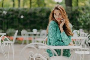 buitenopname van charmante jonge vrouw met lang haar, draagt polka dot groen shirt, zit aan tafel in het terras, heeft een prettig gesprek via moderne smartphone, heeft een dromerige uitdrukking. mensen en levensstijl foto