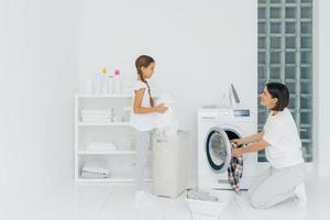drukke brunette vrouw laadt de wasmachine met vuile kleren, haar dochtertje helpt, staat in de buurt van de mand en sorteert de was. gelukkige moeder en kind in de wasruimte. huishoudelijk werk concept. foto