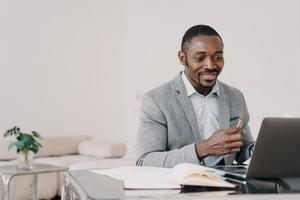 Afro-Amerikaanse zakenman die e-mail leest met goed nieuws op laptop op kantoor, glimlachend foto