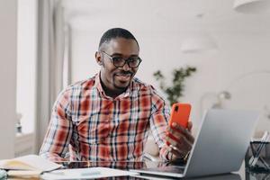 gelukkige afro-man heeft een videogesprek op de telefoon. man blijft thuis in quarantaine en werkt op afstand op laptop. foto